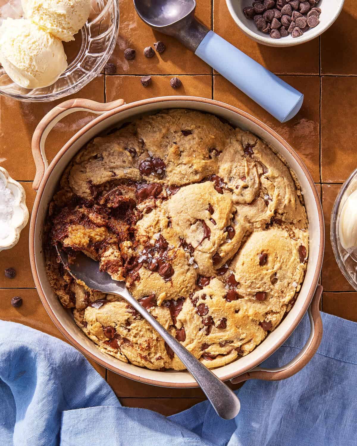 A round baking dish filled with chocolate chip cookie dough dessert, partially scooped out with a spoon. Nearby are bowls of vanilla ice cream, chocolate chips, and a blue napkin on a brown tiled surface.