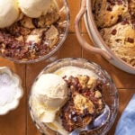 Two glass bowls filled with vanilla ice cream and chocolate chip cookie skillet dessert sit on a tiled surface. A large pan of the cookie dessert and a small bowl of flaky sea salt are nearby. A spoon rests in one bowl.