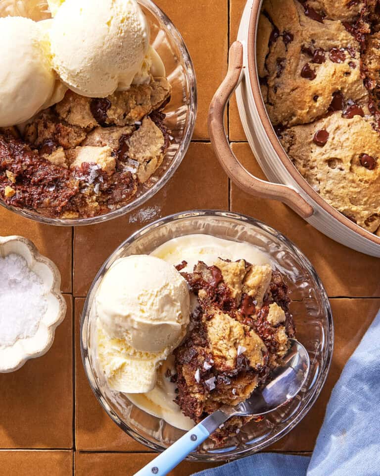 Two glass bowls filled with vanilla ice cream and chocolate chip cookie skillet dessert sit on a tiled surface. A large pan of the cookie dessert and a small bowl of flaky sea salt are nearby. A spoon rests in one bowl.