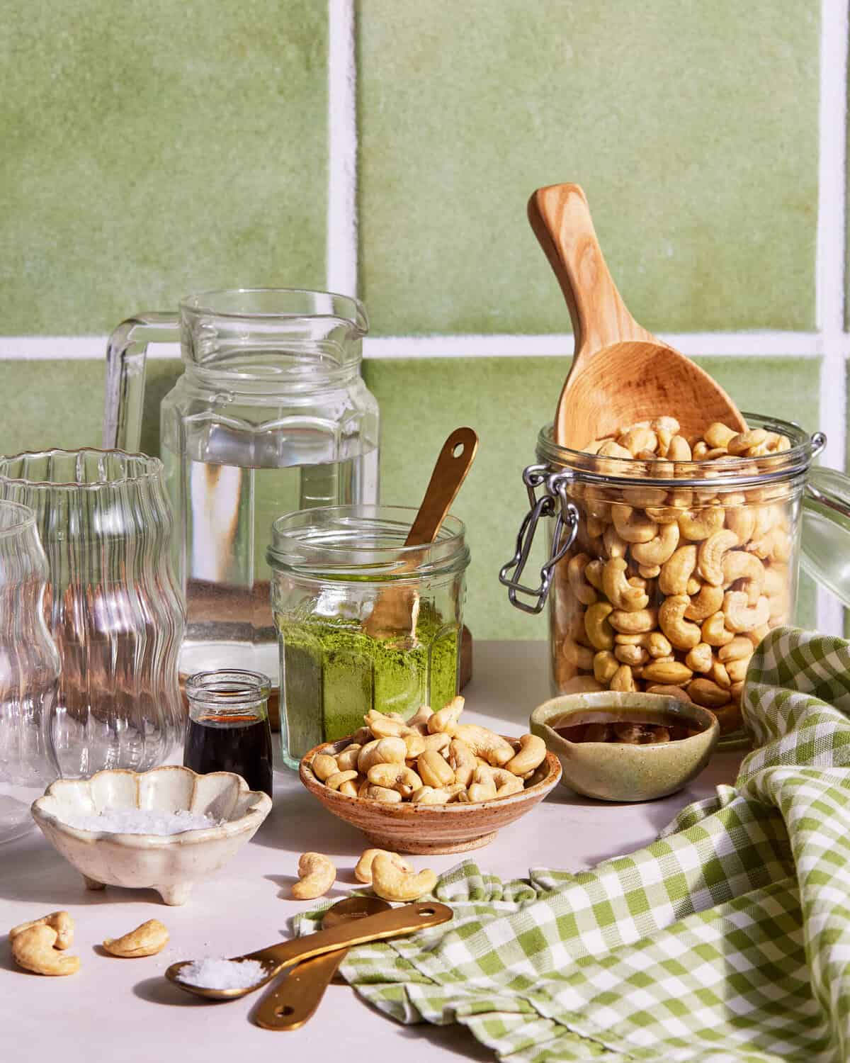 A countertop scene with jars of cashews, a glass pitcher of water, a jar of green powder, a small bowl of salt, a dish of dark liquid, empty glasses, and a green checkered cloth against a tiled wall.
