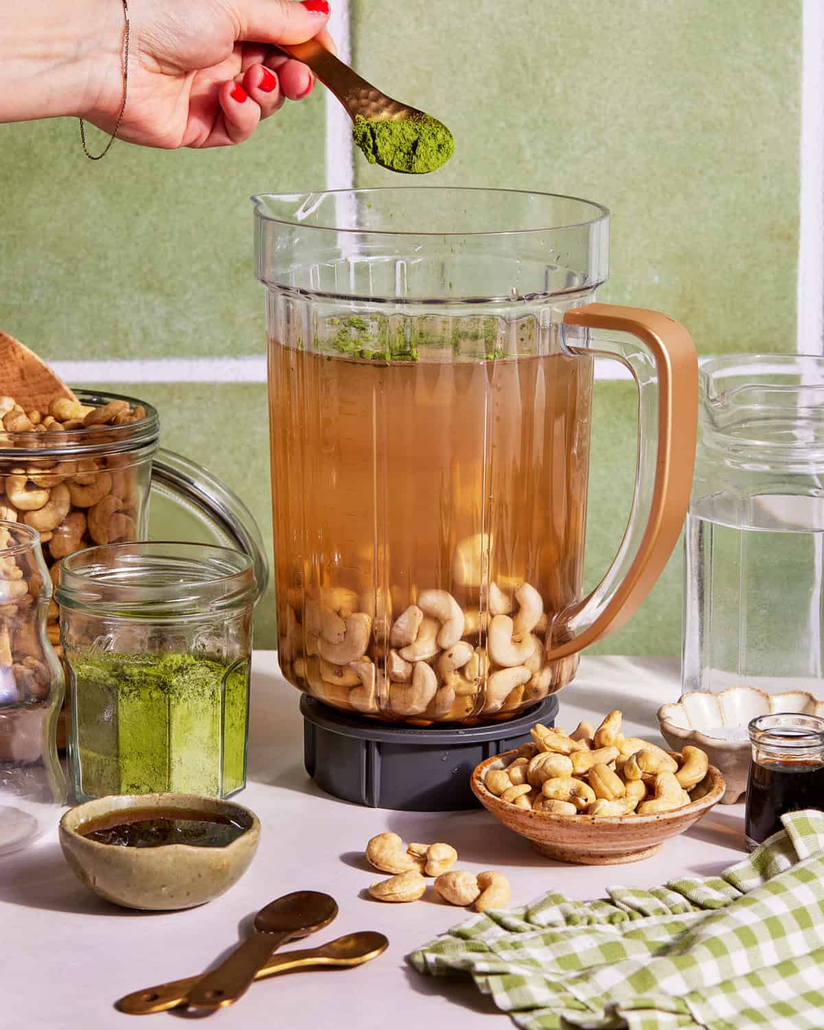 A hand sprinkles green powder into a blender filled with water and cashews. Surrounding the blender are jars of cashews, green powder, a bowl of cashews, and a small cup with dark liquid on a kitchen counter.