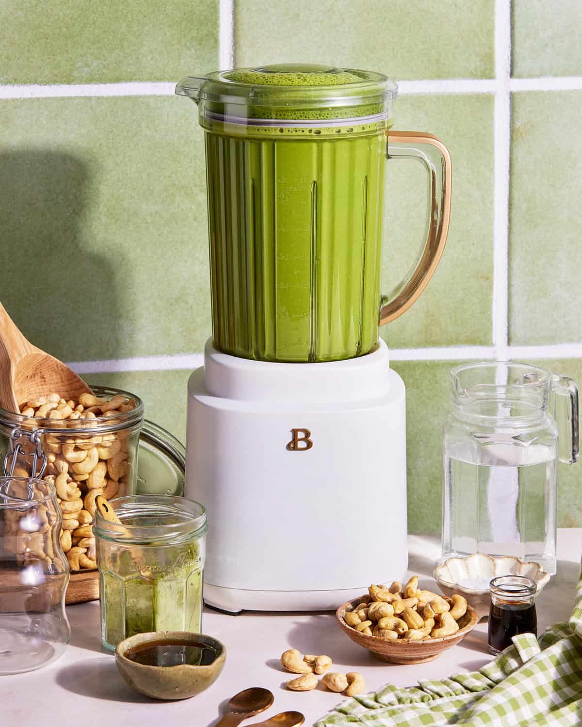 A white blender filled with green smoothie sits on a counter, surrounded by cashews, glass jars, a small bowl, a jug of water, and a green checked cloth, against a light green tiled wall.