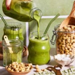 A hand pours a green smoothie from a glass bottle into a glass with a green straw. Cashews, a jar of cashew nuts, wooden utensils, and a green checkered cloth are on the counter amid tiled green walls.