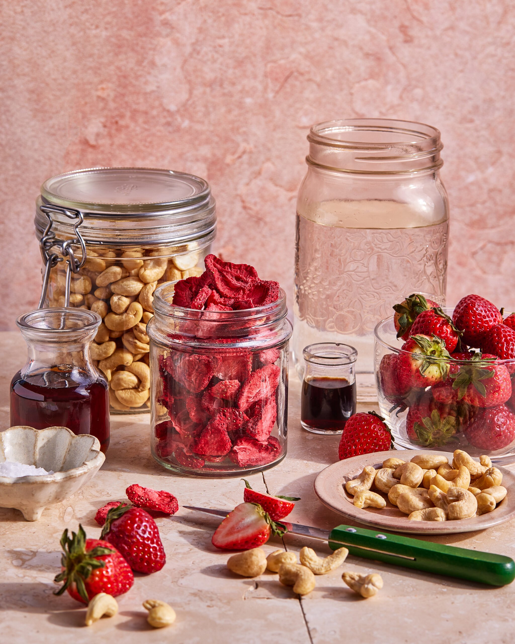 A variety of ingredients, including jars of cashews and freeze-dried strawberries, fresh strawberries, a small bottle of vanilla extract, a jar of maple syrup, a large jar of water, and a green-handled knife on a pink surface.