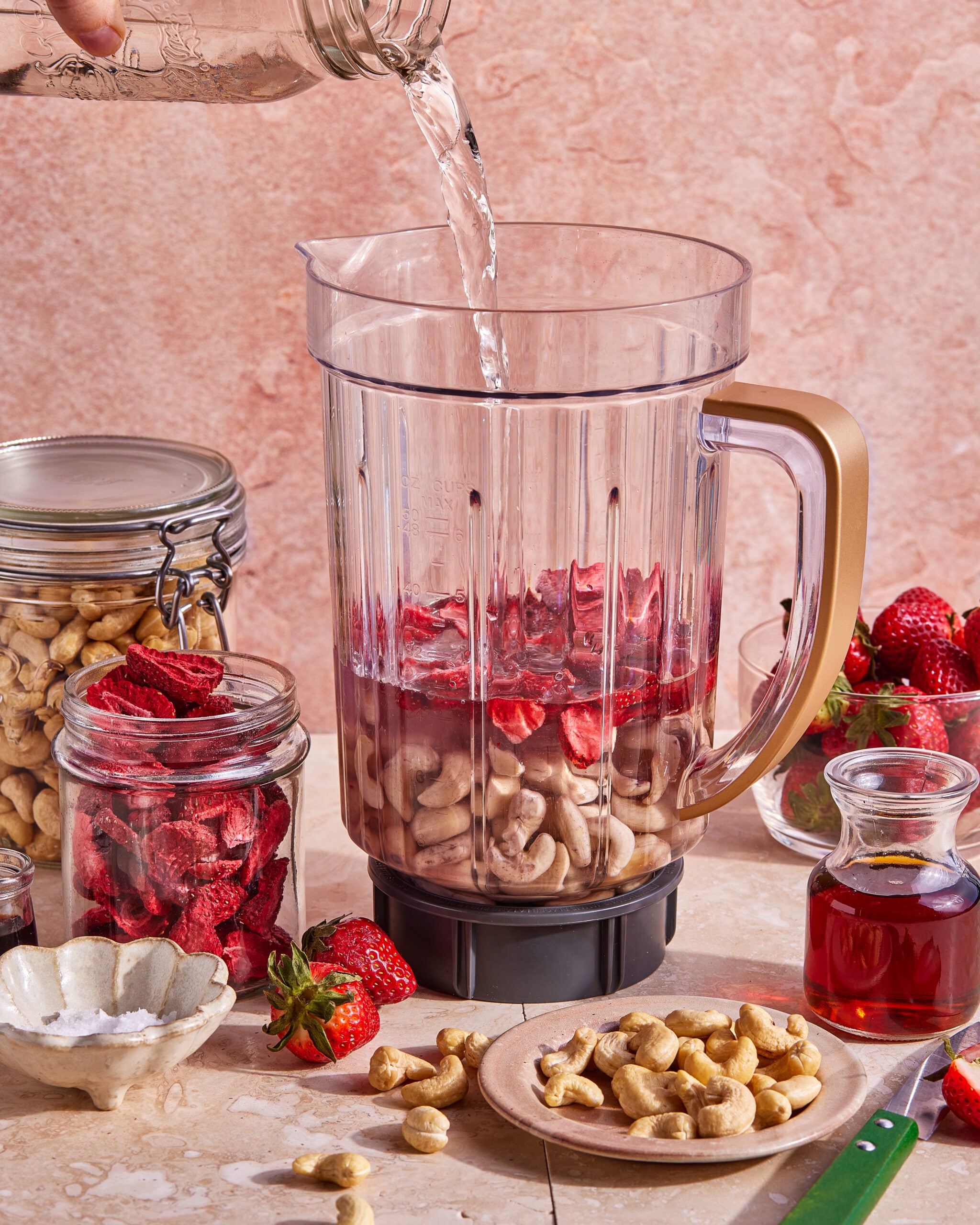 A blender jar filled with cashews and dried strawberries is being filled with water. Surrounding the blender are jars of cashews and dried strawberries, fresh strawberries, a bowl of salt, a knife, and a bottle of syrup.