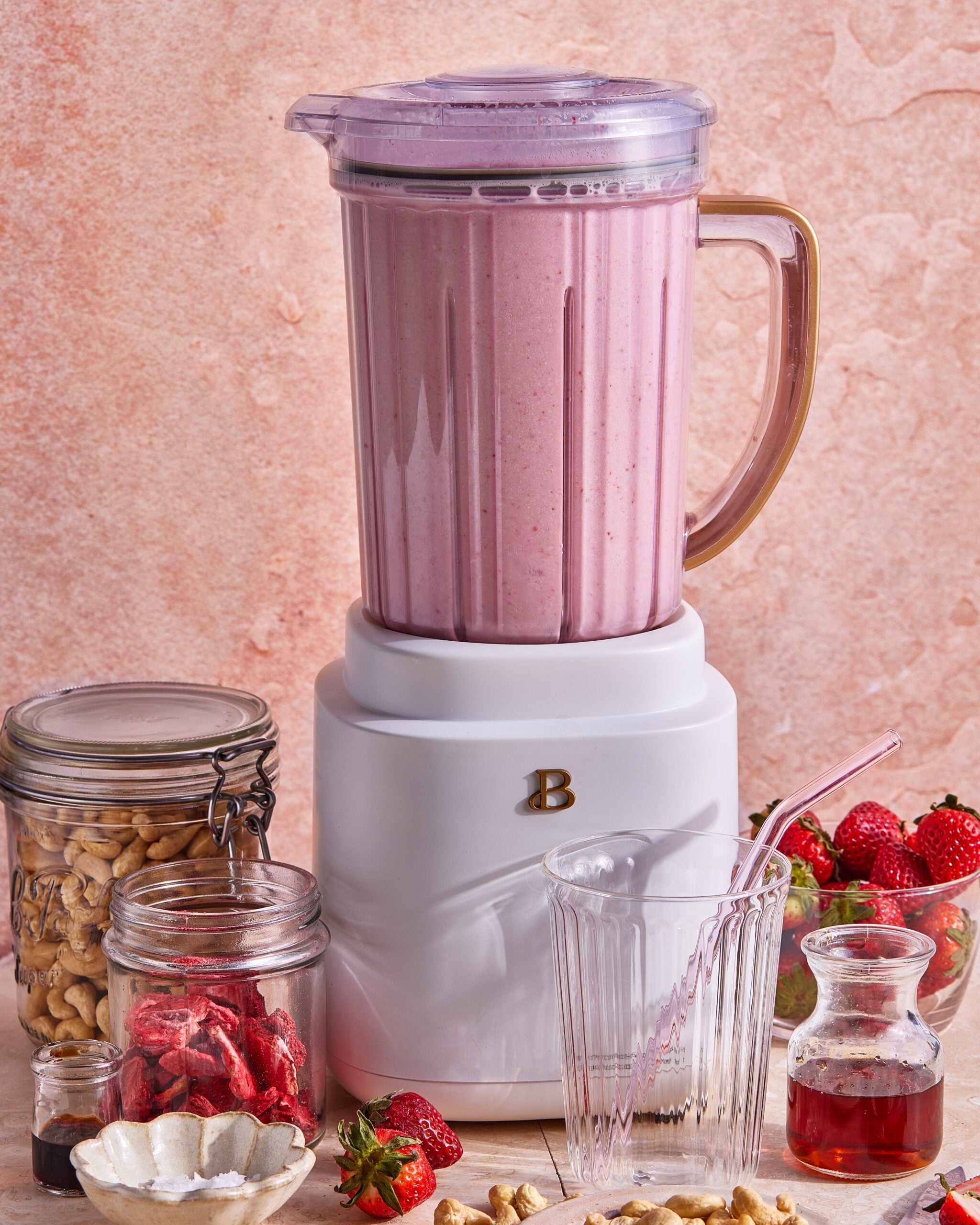 A blender filled with pink smoothie sits on a counter, surrounded by jars of cashews and dried strawberries, fresh strawberries, a glass, and a small glass pitcher with red liquid.