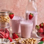 Two glasses of pink strawberry smoothie, each with a strawberry on the rim and a glass straw, are surrounded by fresh and dried strawberries, cashews, and jars of ingredients against a pink background.