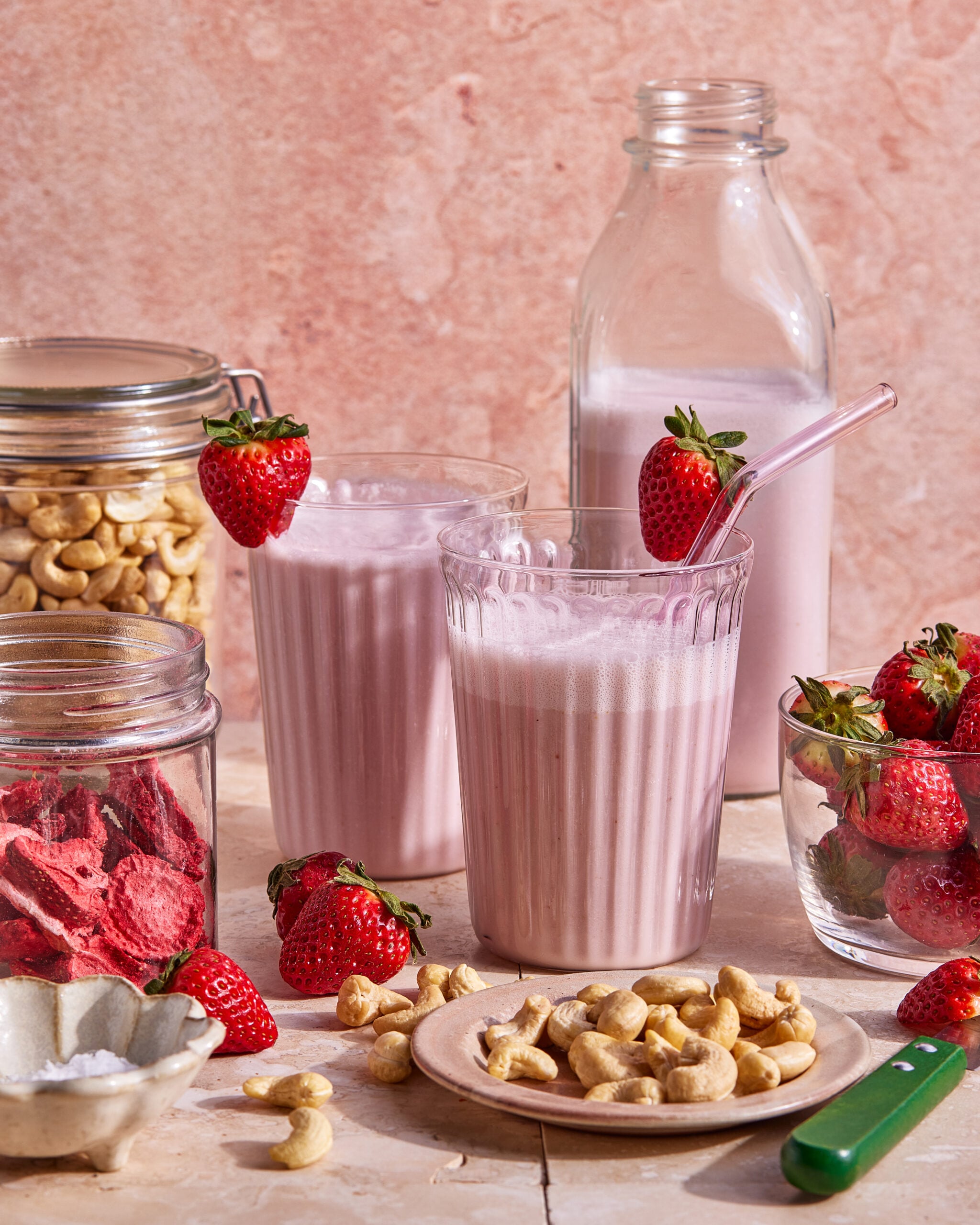 Two glasses of pink strawberry smoothie, each with a strawberry on the rim and a glass straw, are surrounded by fresh and dried strawberries, cashews, and jars of ingredients against a pink background.