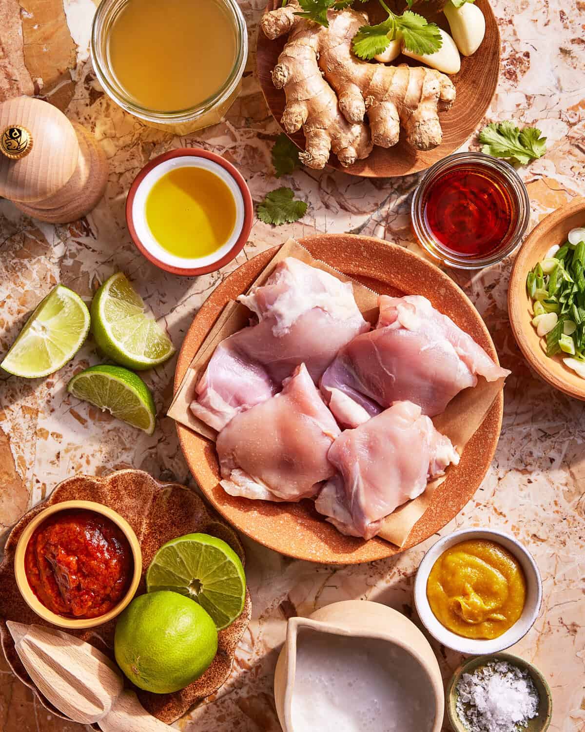 Raw chicken thighs on a plate surrounded by fresh limes, ginger, garlic, green onions, olive oil, broth, chili paste, curry paste, coconut milk, salt, pepper, and red sauce on a marble surface.