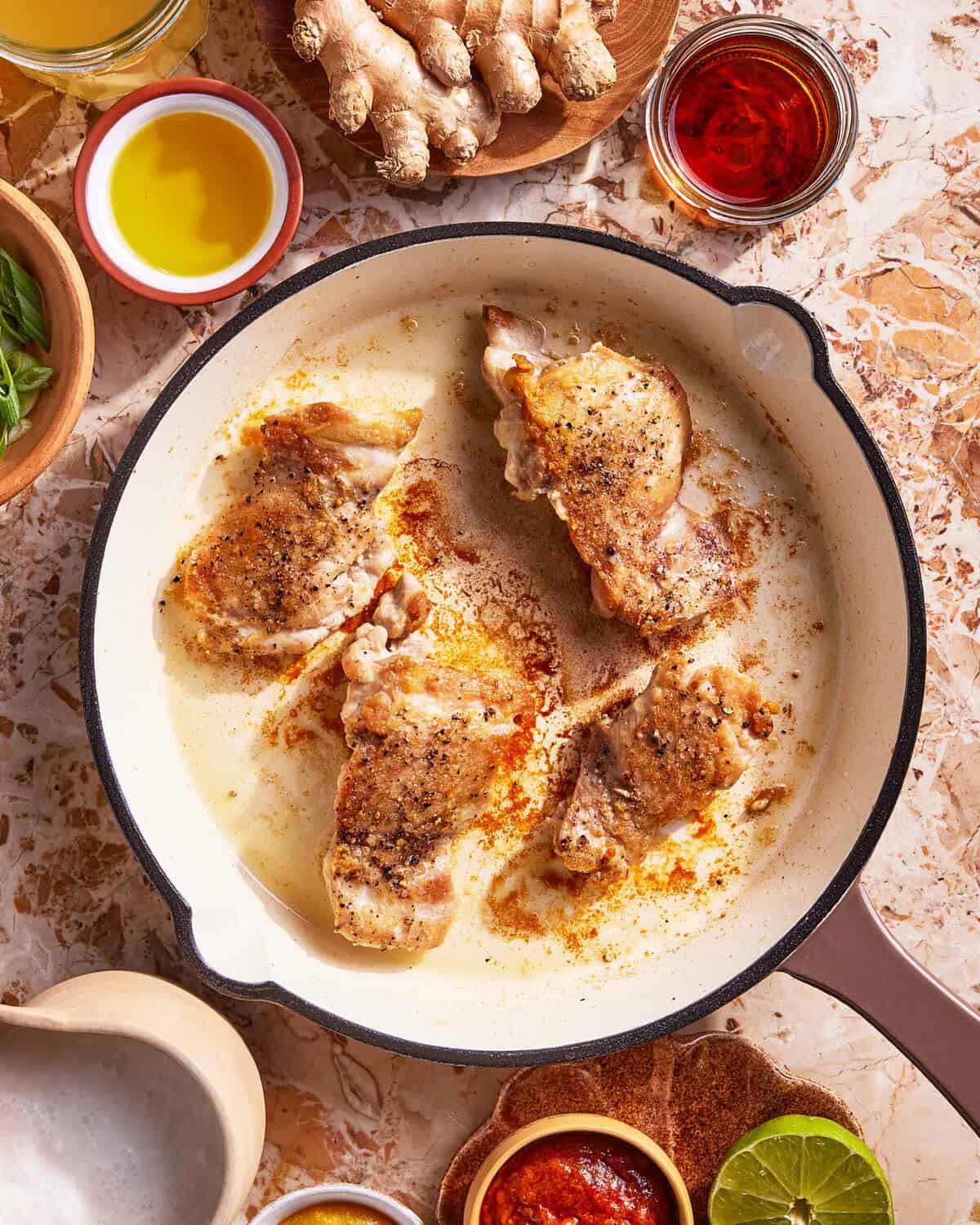 Chicken thighs seasoned with pepper and searing in a white skillet, surrounded by ingredients like ginger, oil, sauces, lime, and herbs on a marbled countertop.