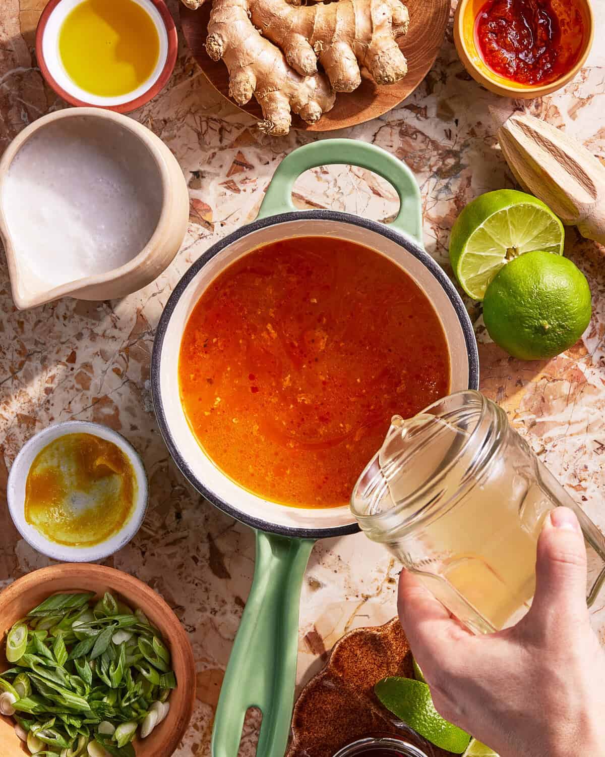 A hand pours liquid from a jar into a saucepan of orange soup, surrounded by ingredients like lime, ginger, scallions, oil, and sauces on a countertop.