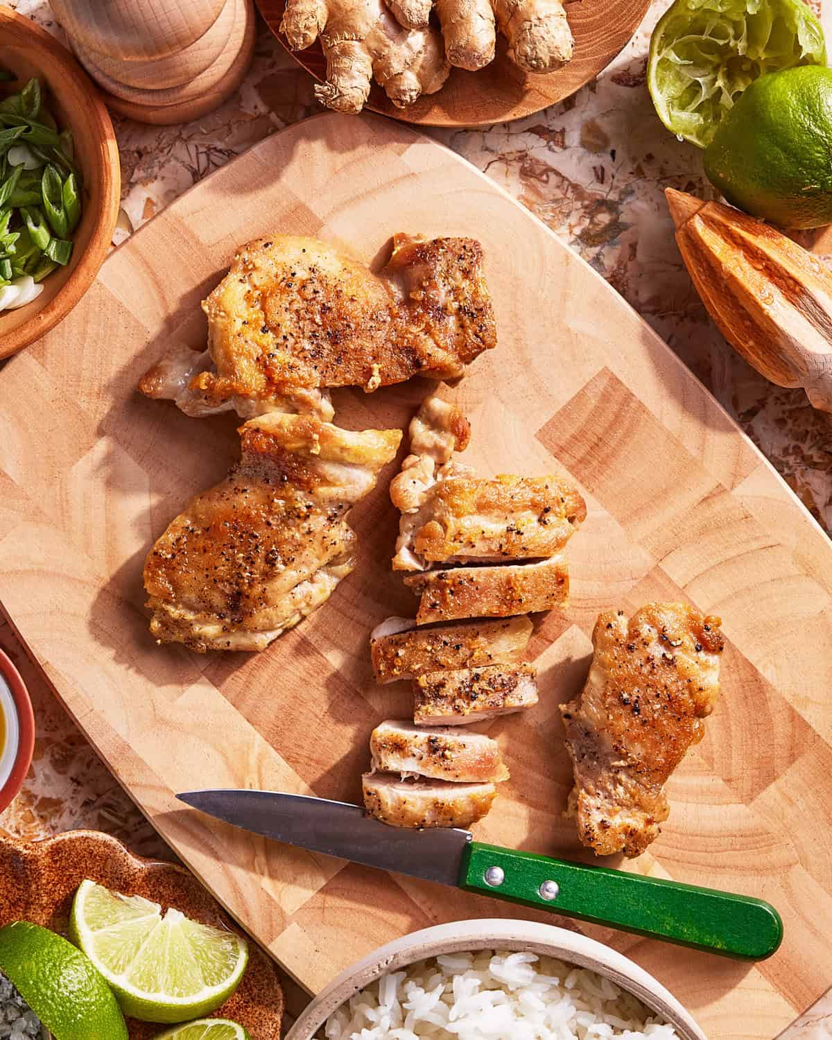 Sliced, cooked chicken thighs on a wooden cutting board with a green-handled knife, surrounded by bowls of rice, lime, green onions, and fresh ginger.