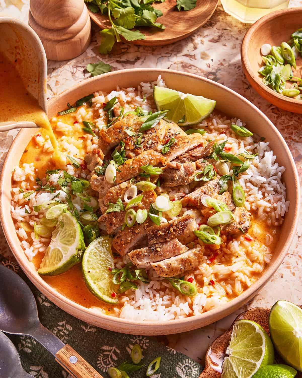 A bowl of rice topped with sliced grilled chicken, creamy orange curry sauce being poured on top, garnished with lime wedges, green onions, and cilantro. Utensils, herbs, and a drink are nearby on the table.
