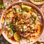 A bowl of rice topped with sliced grilled chicken, chopped green onions, lime wedges, fresh herbs, and a creamy sauce. A spoon rests in the bowl, and extra lime wedges and herbs are nearby.