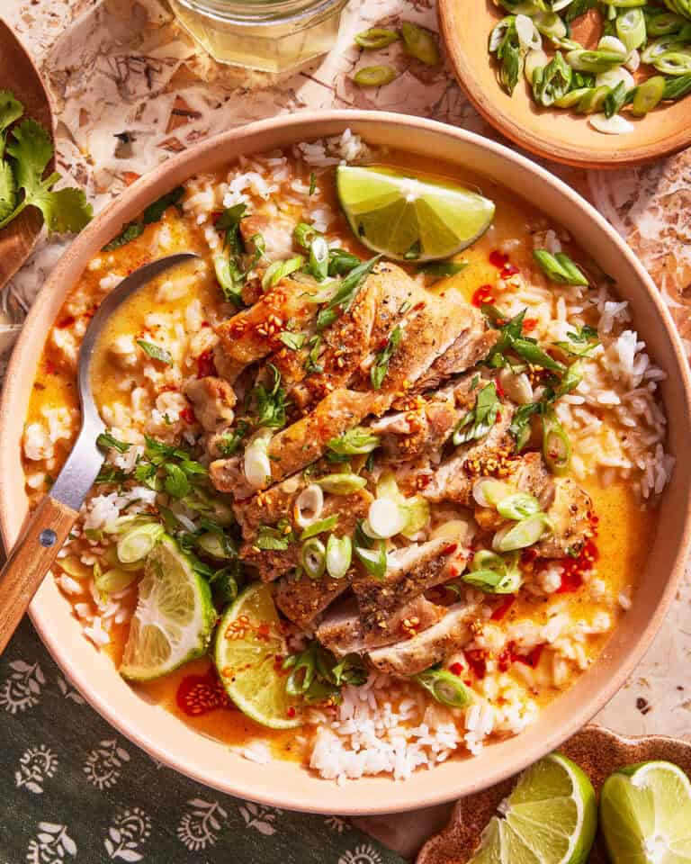 A bowl of rice topped with sliced grilled chicken, chopped green onions, lime wedges, fresh herbs, and a creamy sauce. A spoon rests in the bowl, and extra lime wedges and herbs are nearby.