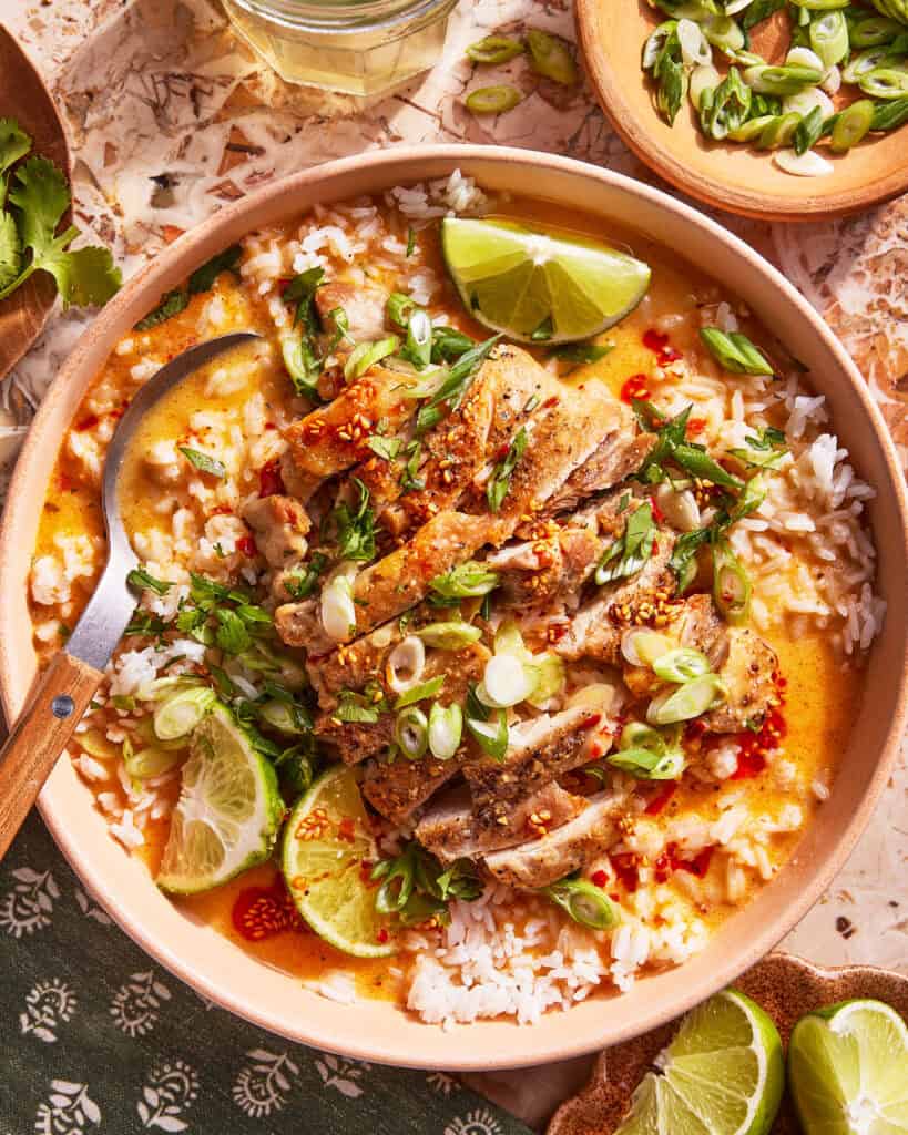 A bowl of rice topped with sliced grilled chicken, chopped green onions, lime wedges, fresh herbs, and a creamy sauce. A spoon rests in the bowl, and extra lime wedges and herbs are nearby.