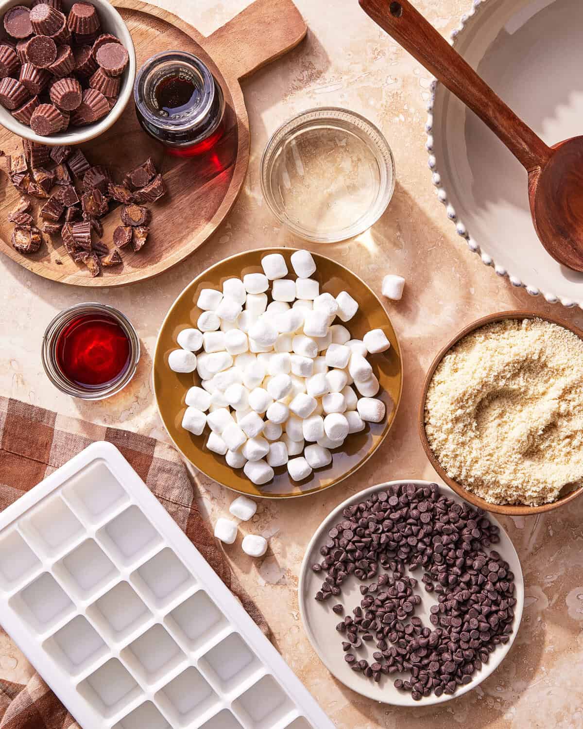 Overhead view of baking ingredients: mini marshmallows, chocolate chips, chopped peanut butter cups, almond flour, two glasses of liquid (clear and red), in bowls on a countertop beside an empty ice cube tray and pie dish.