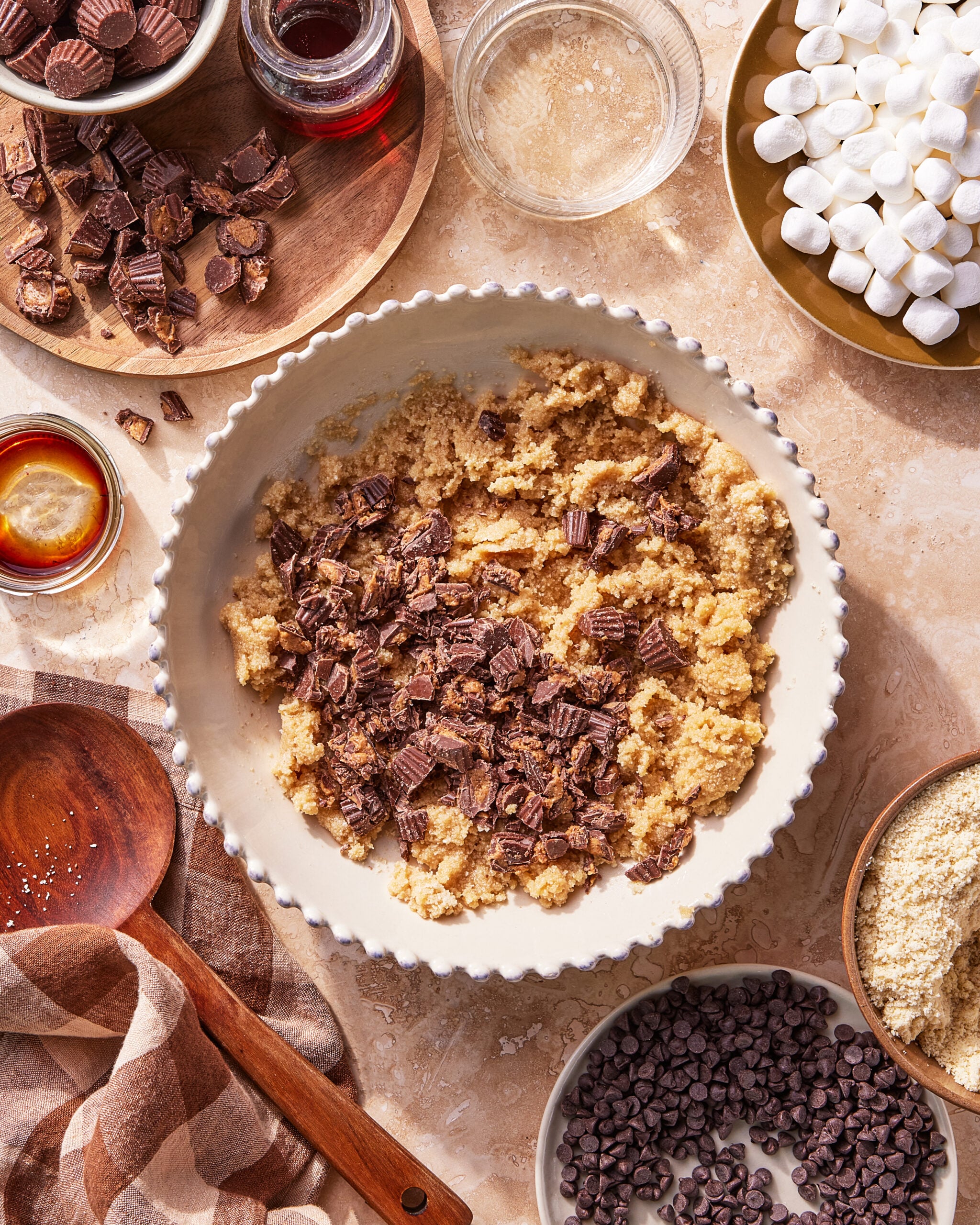 A large bowl of cookie dough topped with chopped peanut butter cups is surrounded by bowls of mini marshmallows, chocolate chips, chopped peanut butter cups, and various baking ingredients on a beige surface.