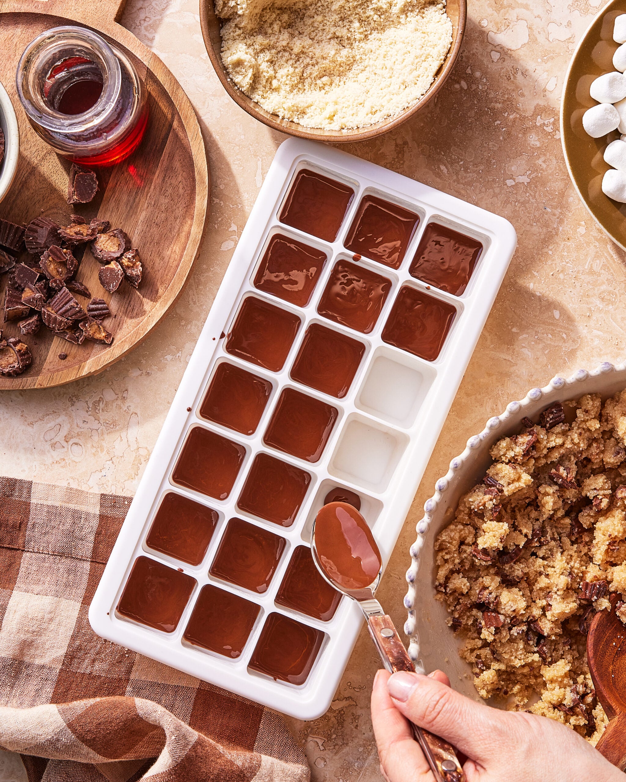 A hand fills an ice cube tray with melted chocolate using a spoon. Surrounding the tray are bowls of chopped chocolate, almond flour, cookie crumbs, marshmallows, and a jar of red liquid on a beige countertop.