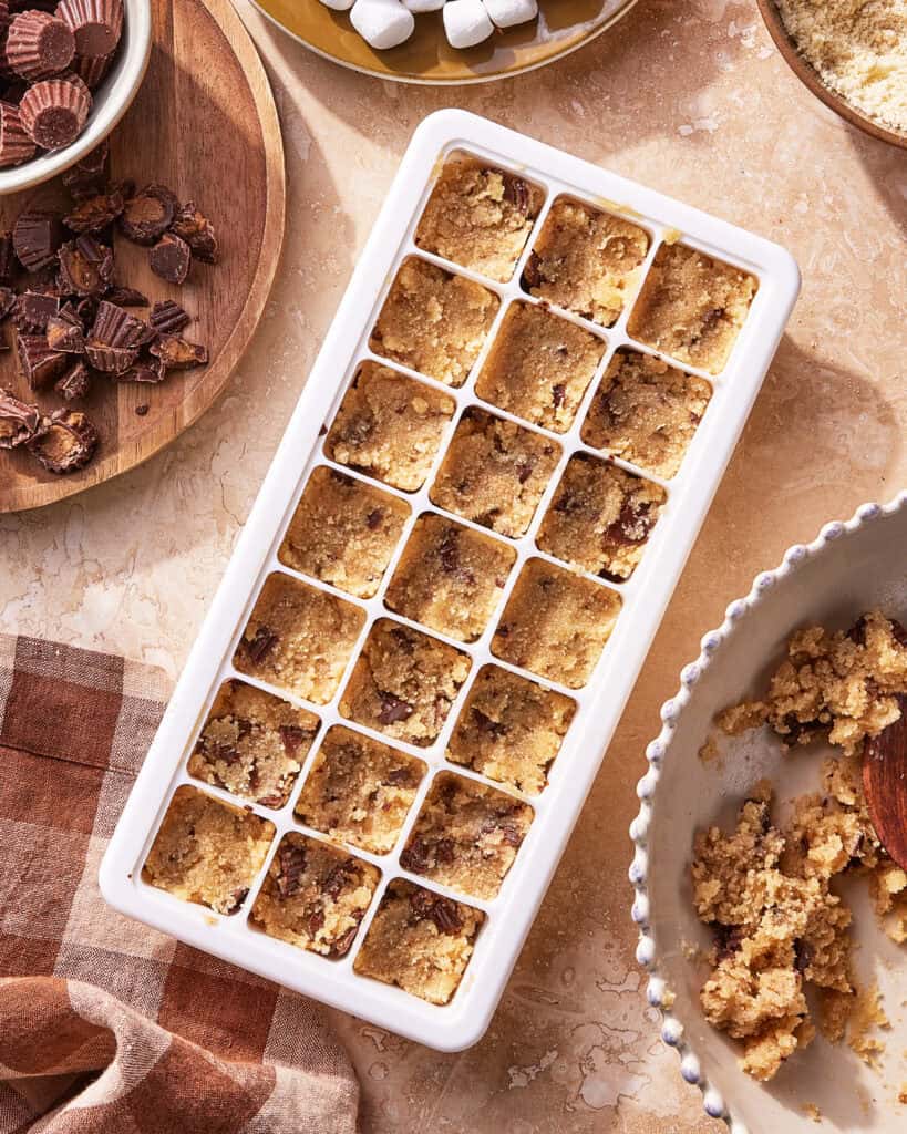 A white ice cube tray filled with chocolate chip cookie dough is on a counter surrounded by bowls of chopped peanut butter cups, marshmallows, and extra dough. A checked cloth napkin is nearby.