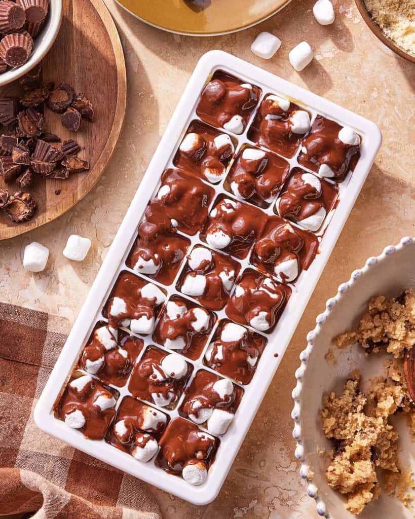 A white ice cube tray filled with melted chocolate and mini marshmallows sits on a tan countertop, surrounded by bowls of chopped peanut butter cups and crumbly brown sugar mixture.