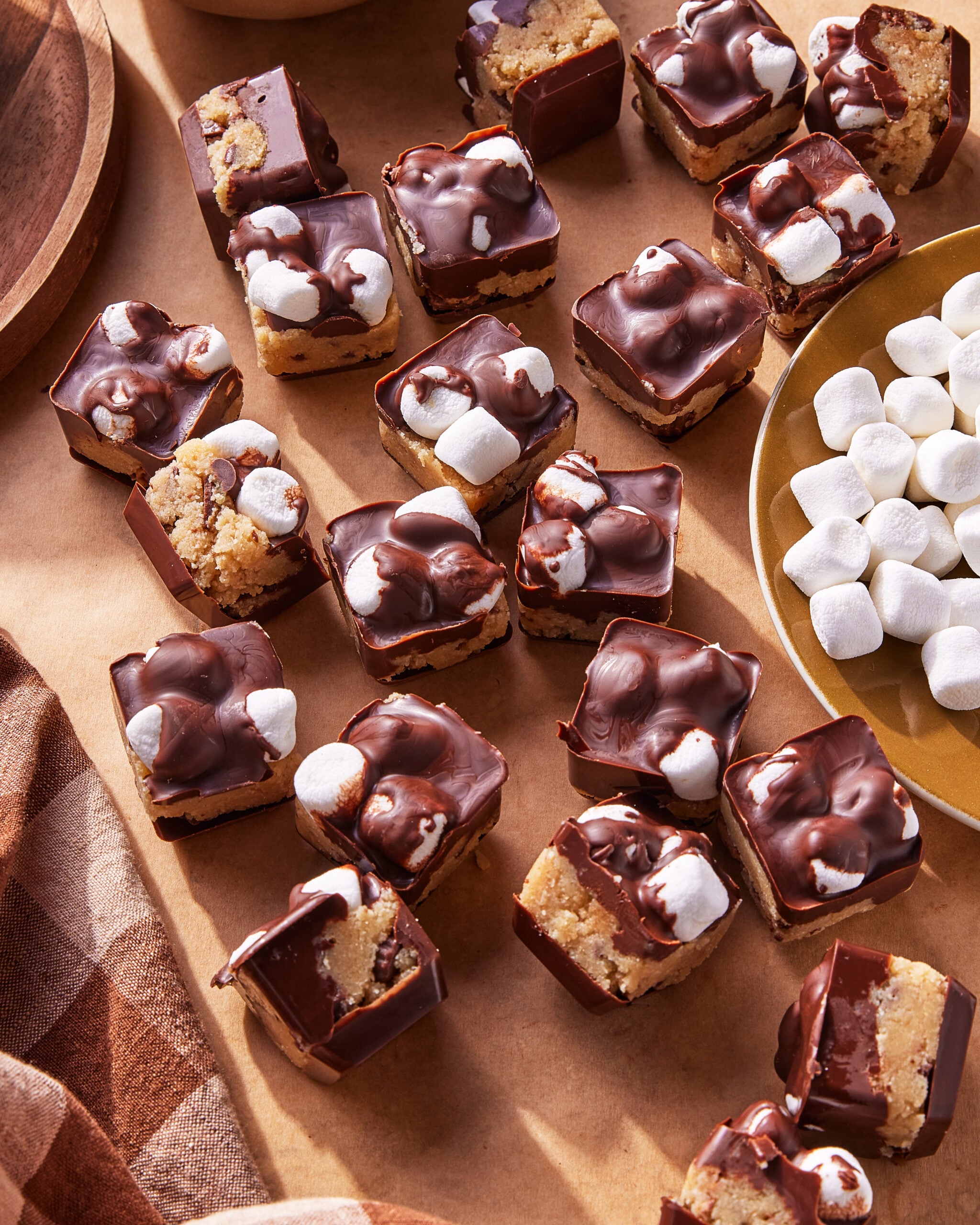 Squares of chocolate-covered dessert topped with mini marshmallows are arranged on a brown surface next to a plate of loose mini marshmallows and a brown-checkered cloth. The treats have a layered appearance.
