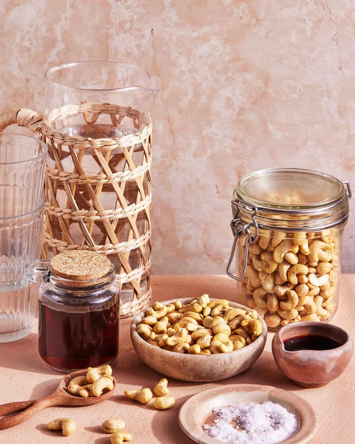 A still life of a glass pitcher with a wicker cover, a glass of water, a jar of cashews, a small bottle of dark liquid, bowls of cashews, a small dish of salt, and a wooden spoon, all on a beige surface.