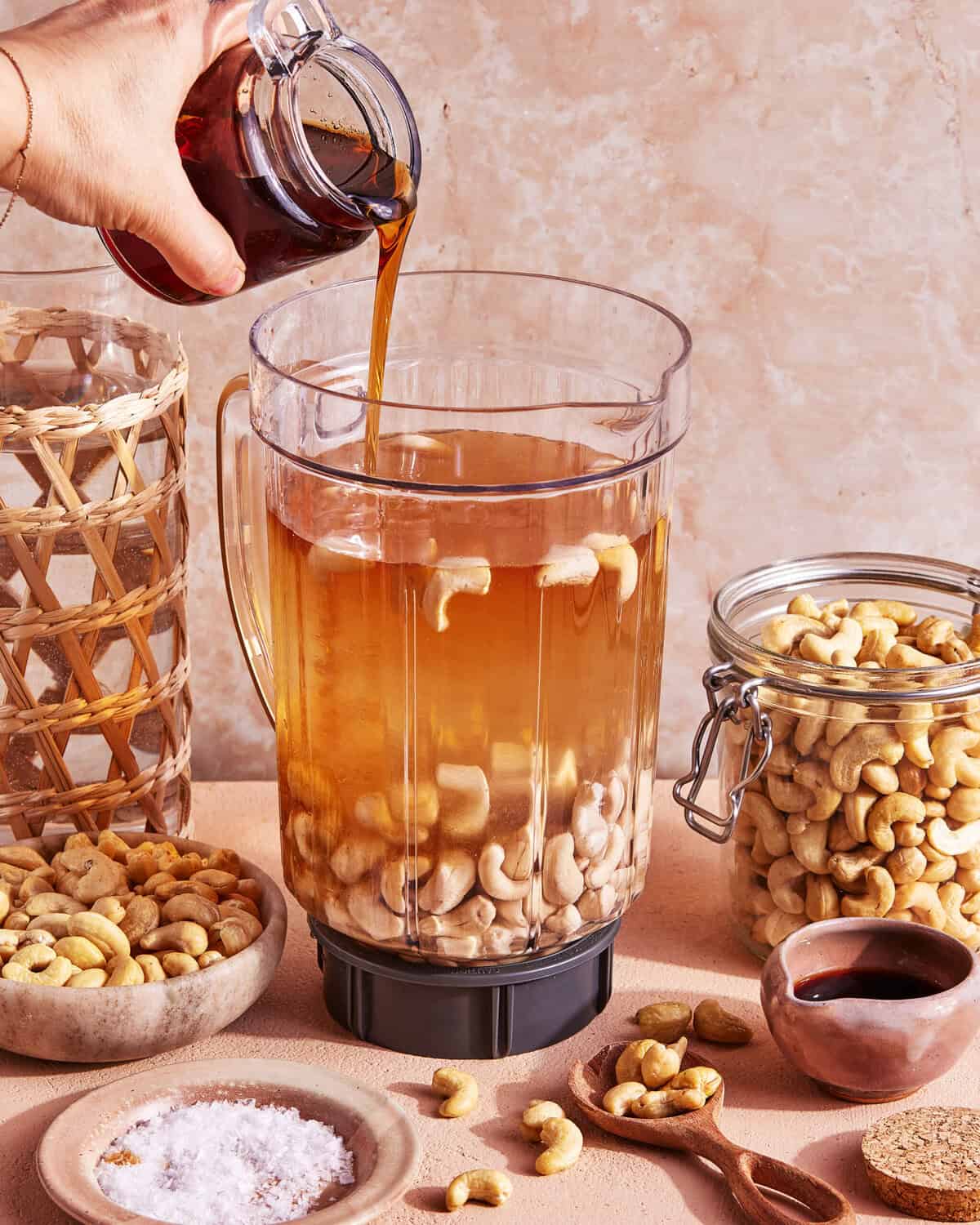 A hand pours a dark liquid, likely coffee, into a blender filled with cashews and water. Surrounding the blender are jars and bowls of cashews, a bowl of salt, a small bowl of brown paste, and a small scoop of powder.