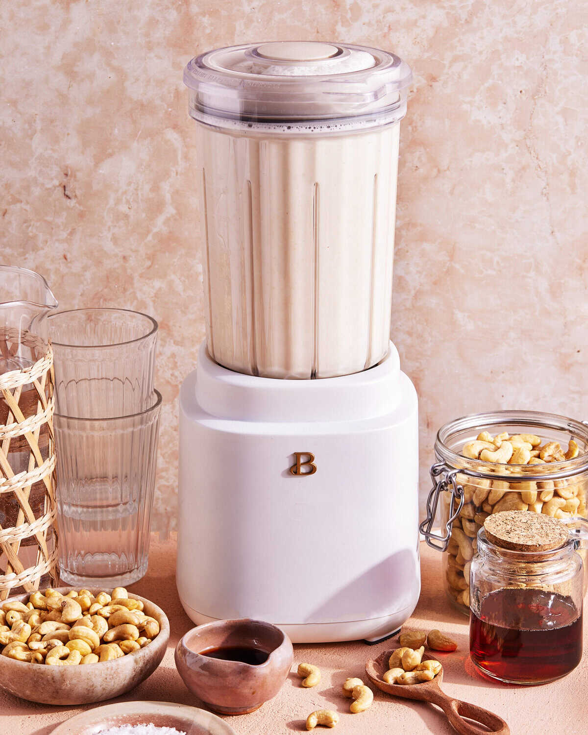 A white blender filled with a creamy mixture sits on a counter surrounded by glassware, a jar of cashews, a small bowl of honey, loose cashews, and a decorative basket.