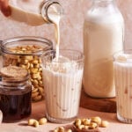 A hand pours creamy cashew milk into a glass filled with ice. Nearby are jars of cashews and syrup, another glass of cashew milk, a large bottle of cashew milk, and scattered cashews on a light surface.