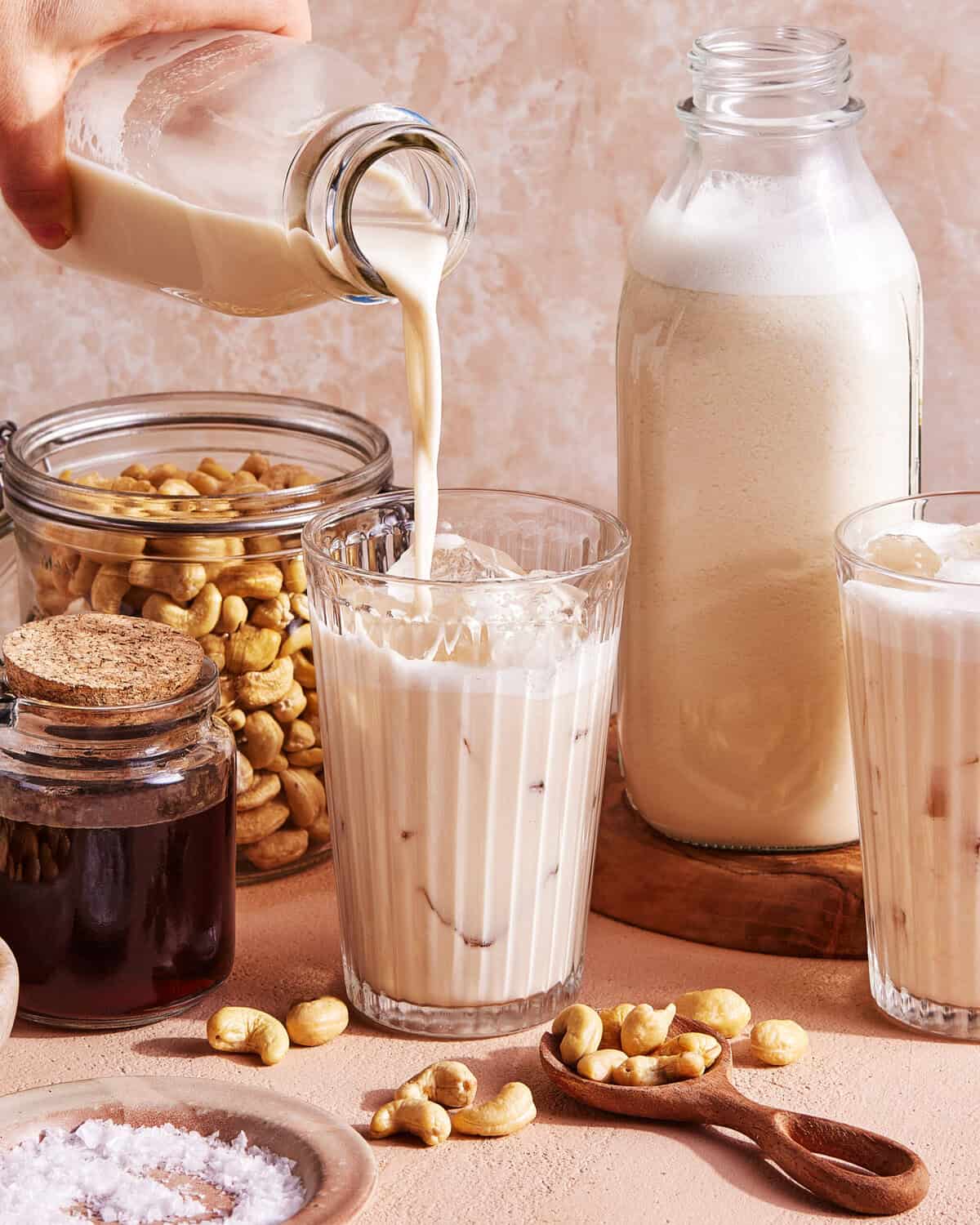 A hand pours creamy cashew milk into a glass filled with ice. Nearby are jars of cashews and syrup, another glass of cashew milk, a large bottle of cashew milk, and scattered cashews on a light surface.