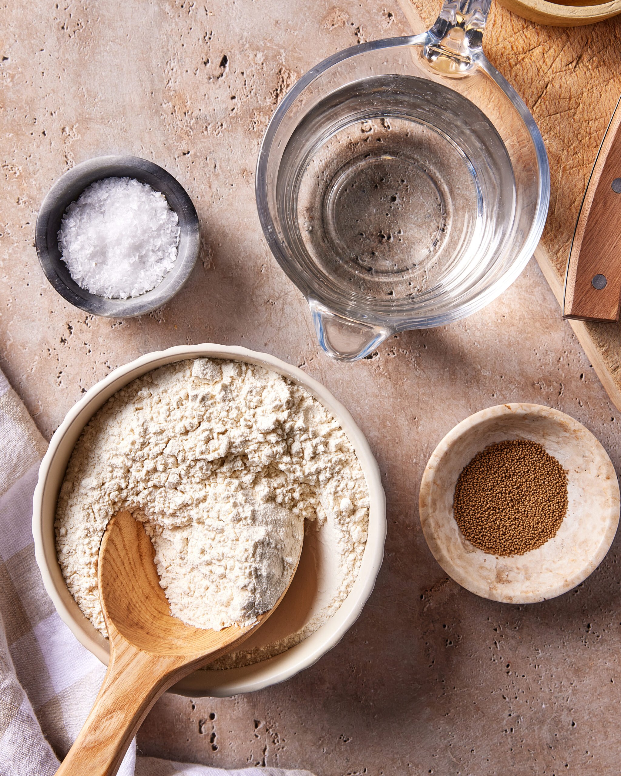 A bowl of flour with a wooden spoon, a glass measuring cup of water, a small bowl of salt, and a small bowl of dry yeast are arranged on a textured surface next to a wooden cutting board.