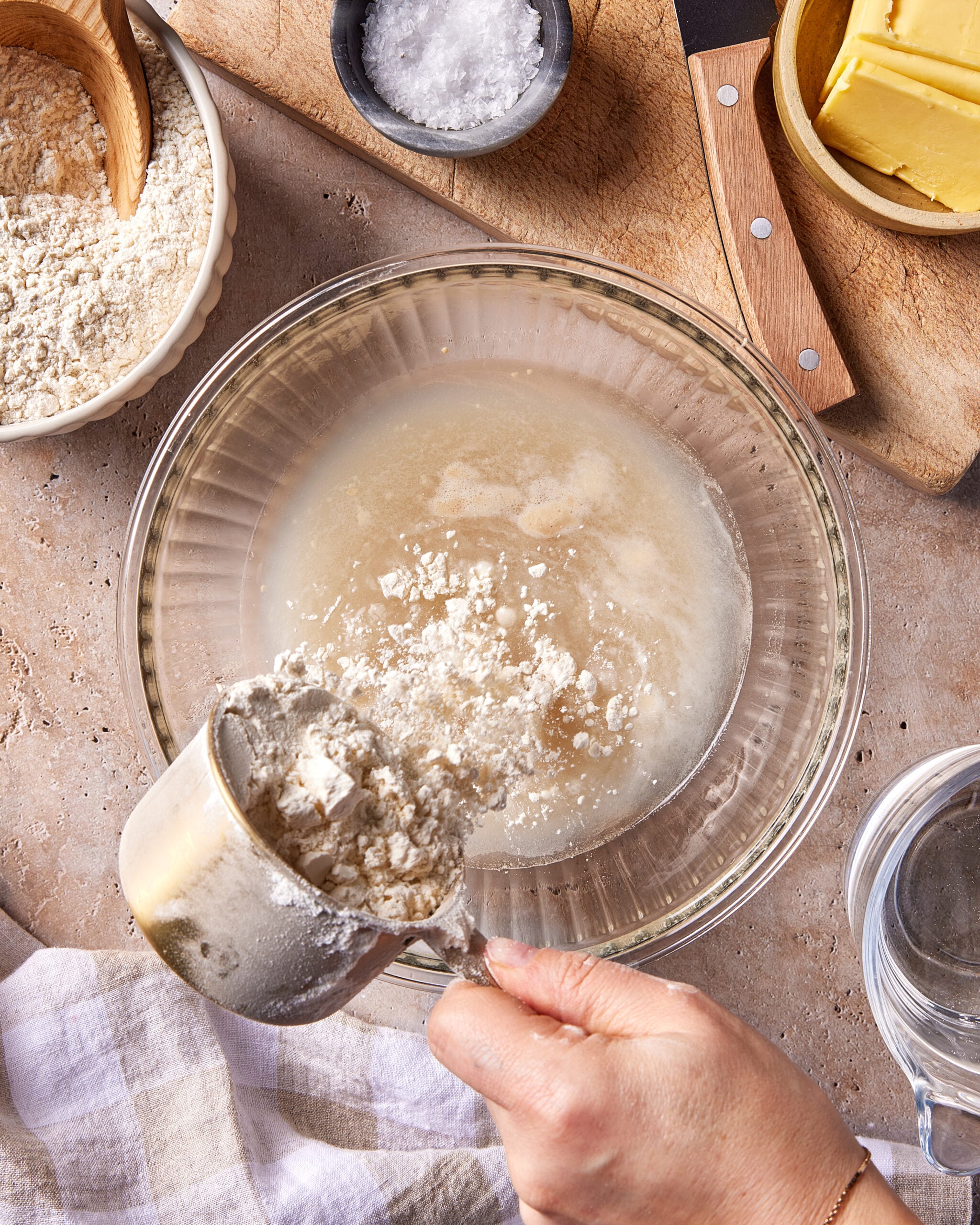 A hand pours flour from a metal measuring cup into a glass bowl filled with water and yeast. Surrounding the bowl are ingredients: flour, butter, salt, and a small pitcher of water on a kitchen counter.