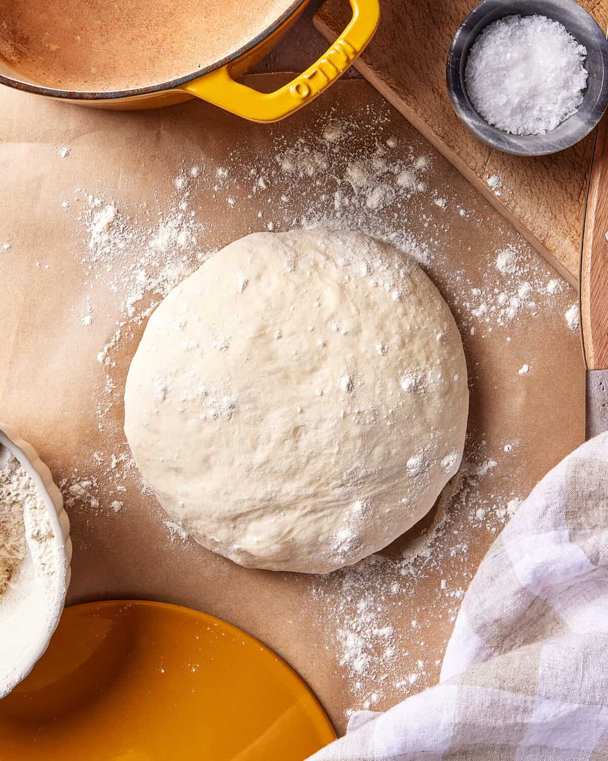 A ball of pizza dough dusted with flour rests on parchment paper, surrounded by a bowl of flour, a yellow dish, a cloth, and a small bowl of salt.