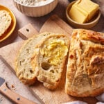 A loaf of rustic bread is partially sliced on a wooden cutting board, with two buttered slices in front. Nearby are a small bowl of flour, a plate of butter, a dish of salt, a bread knife, and a bread roll.
