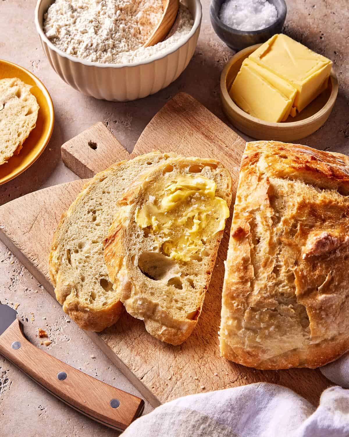 A loaf of rustic bread is partially sliced on a wooden cutting board, with two buttered slices in front. Nearby are a small bowl of flour, a plate of butter, a dish of salt, a bread knife, and a bread roll.