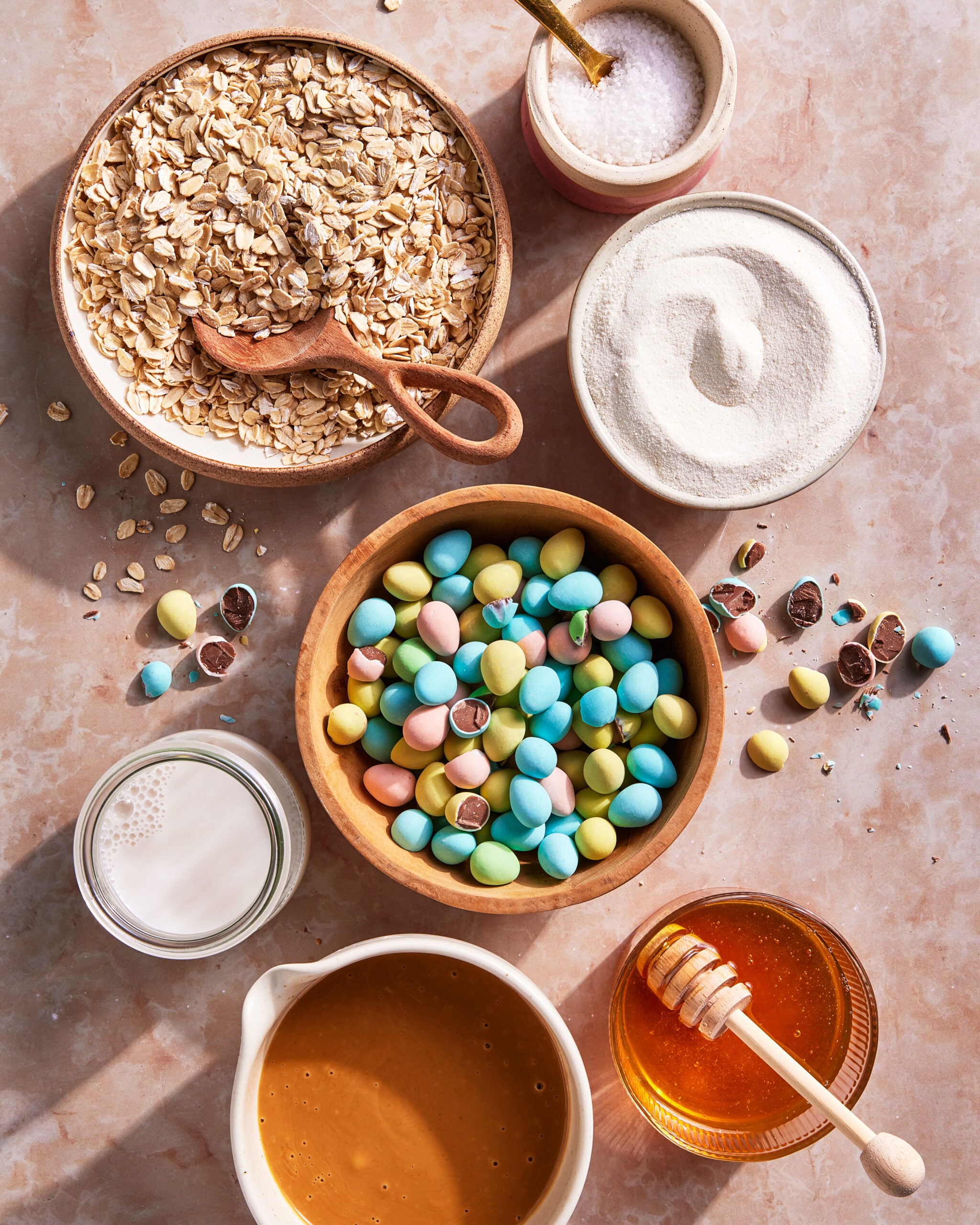 A top-down view of bowls containing oats, sugar, pastel candy-coated chocolates, a glass of milk, a bowl of honey with a honey dipper, caramel sauce, and a small bowl of salt on a light marble surface.