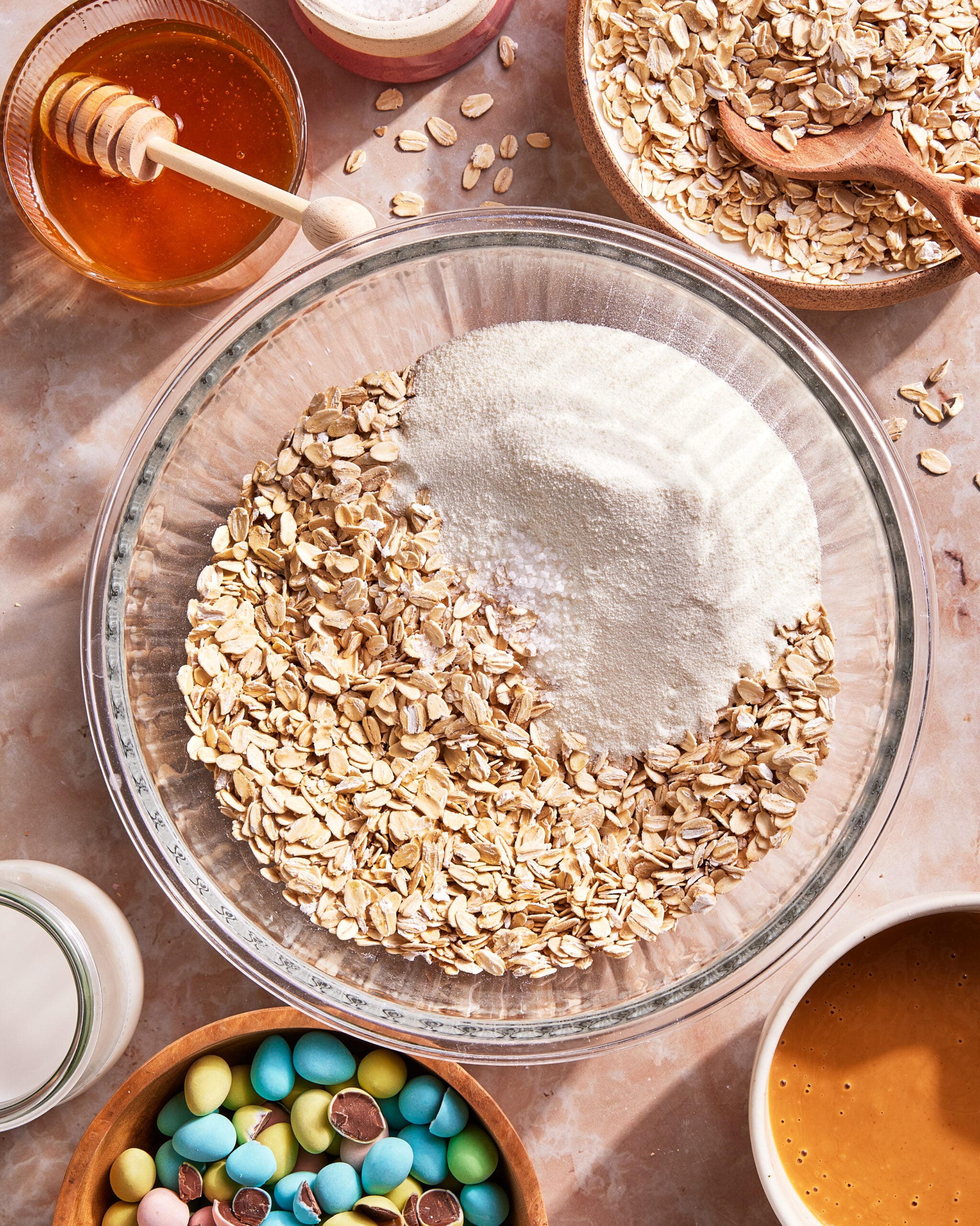 A glass bowl with oats and flour on a marble surface, surrounded by honey, a bowl of oats with a wooden spoon, a bowl of mini chocolate eggs, a bottle of milk, and a bowl of peanut butter.