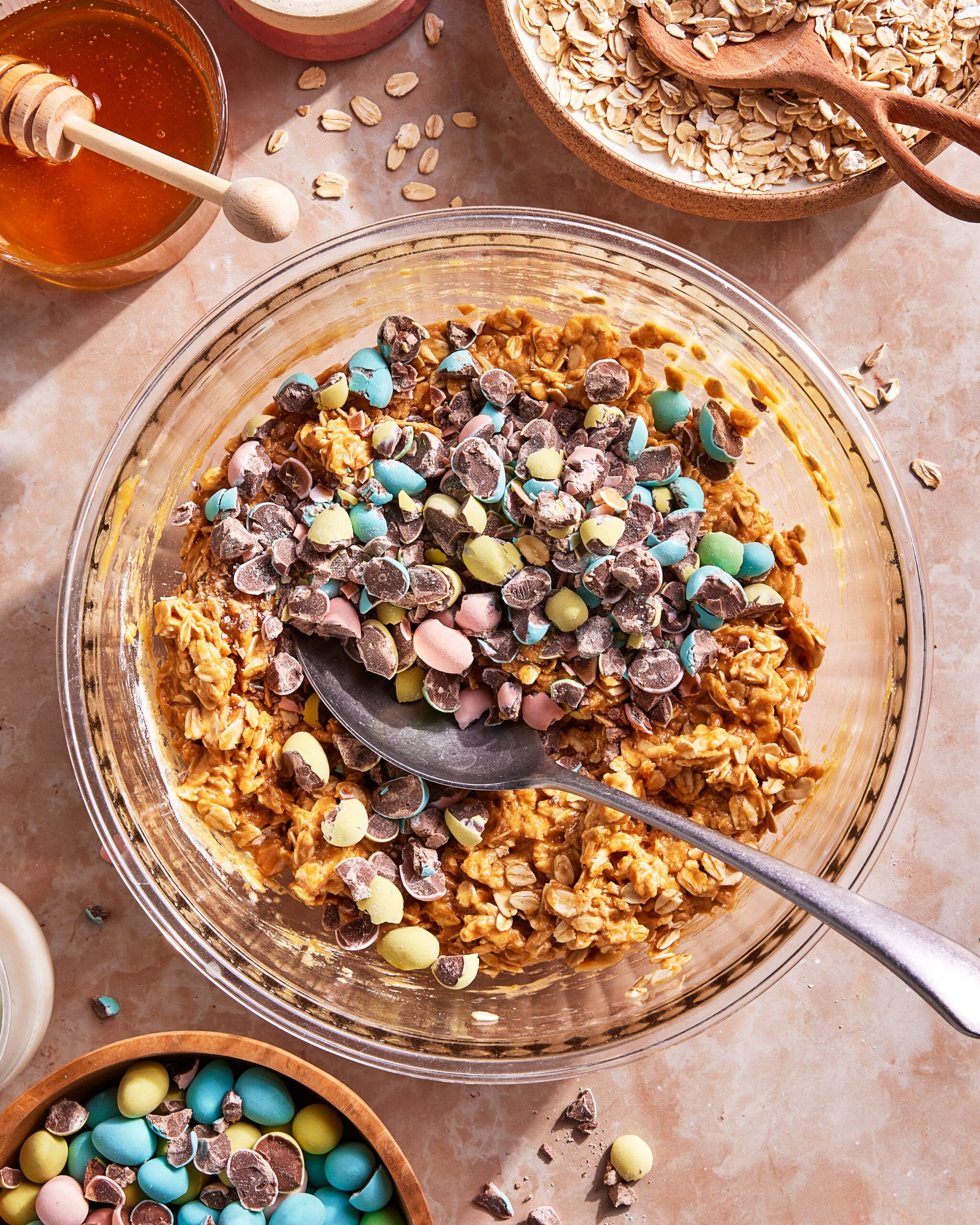 A bowl of oatmeal mixture topped with chopped pastel-colored candy-coated chocolates, surrounded by bowls of oats, honey, and more candies on a light countertop. A spoon rests in the oatmeal mixture.