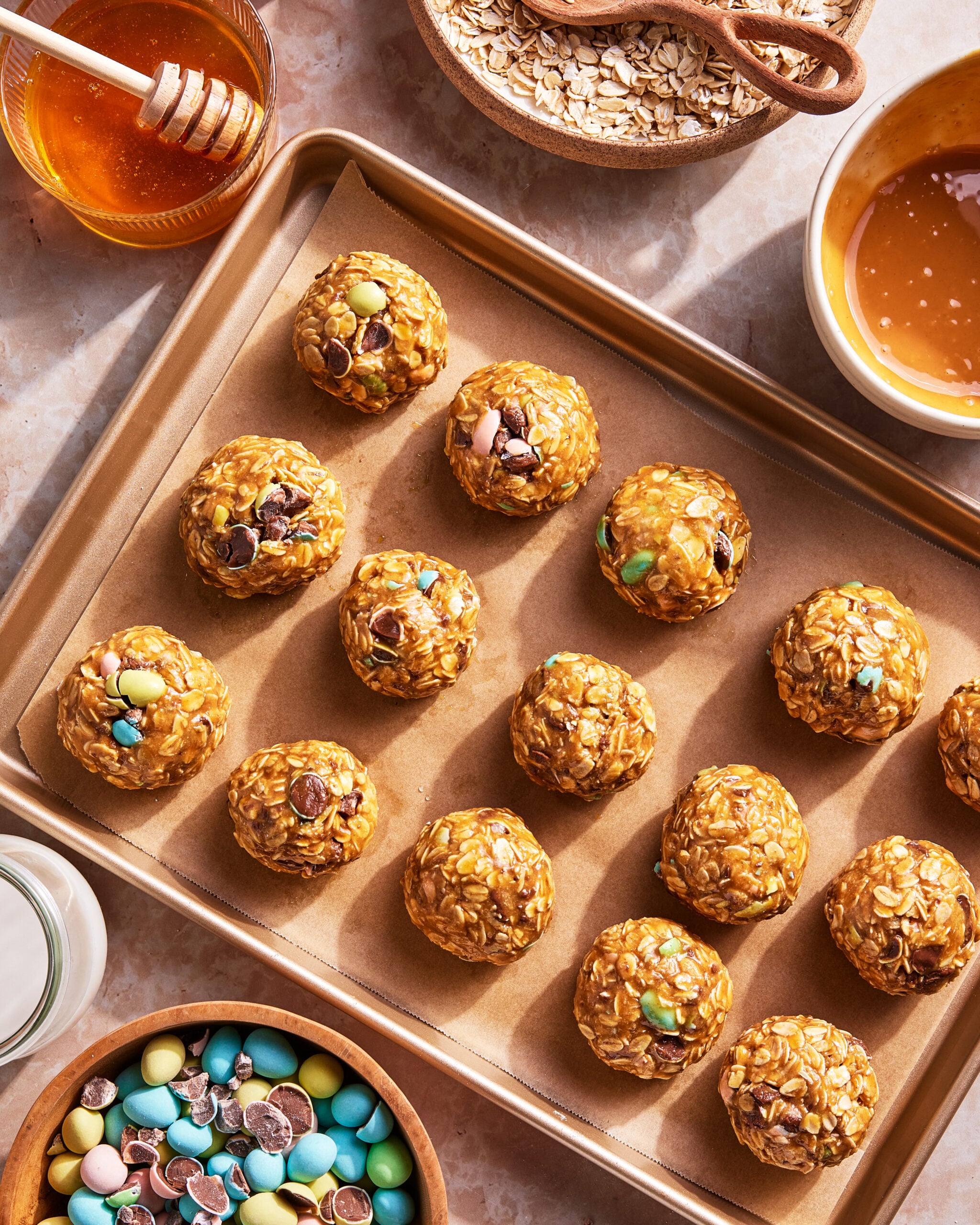 A baking sheet with round oat and candy energy bites, surrounded by bowls of honey, oats, colorful candies, and a glass of milk on a light countertop.