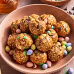 A bowl filled with oat and chocolate chip energy bites, surrounded by pastel-colored candy-coated chocolate eggs. A jar of honey and a wooden honey dipper are in the background.