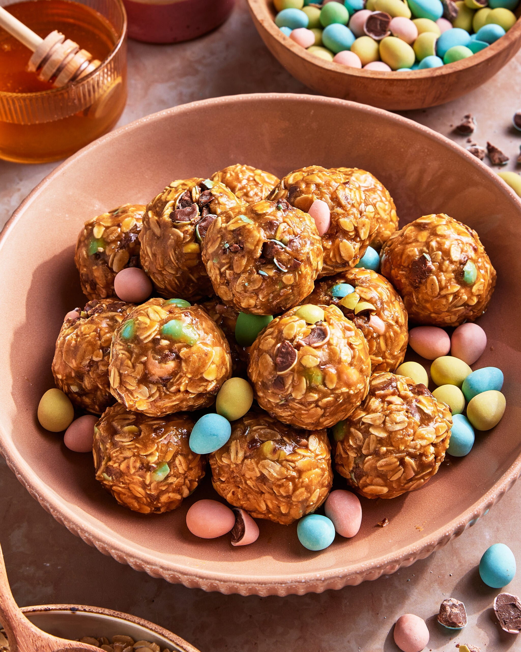 A bowl filled with oat and chocolate chip energy bites, surrounded by pastel-colored candy-coated chocolate eggs. A jar of honey and a wooden honey dipper are in the background.