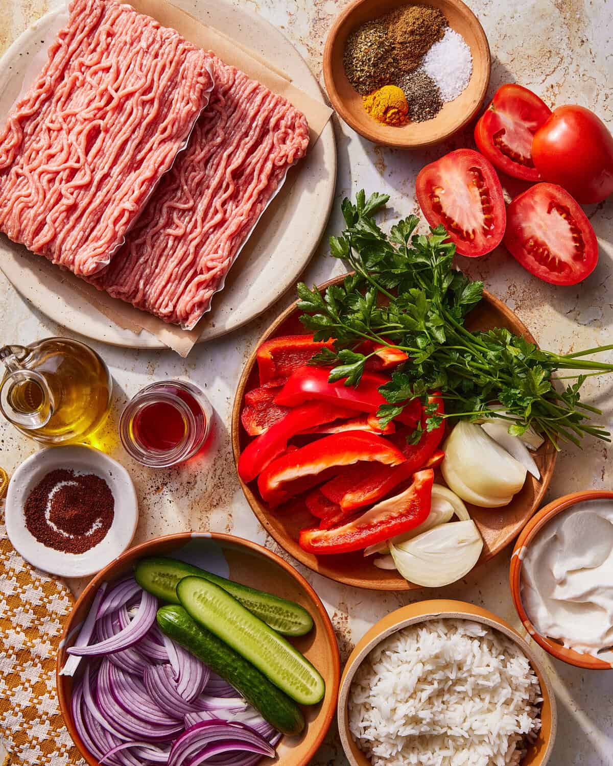 Raw ground meat, spices, salt, fresh tomatoes, sliced red bell pepper, parsley, onion, cucumber, red onion, olive oil, red vinegar, sumac, white rice, and a small bowl of creamy sauce arranged on a counter.