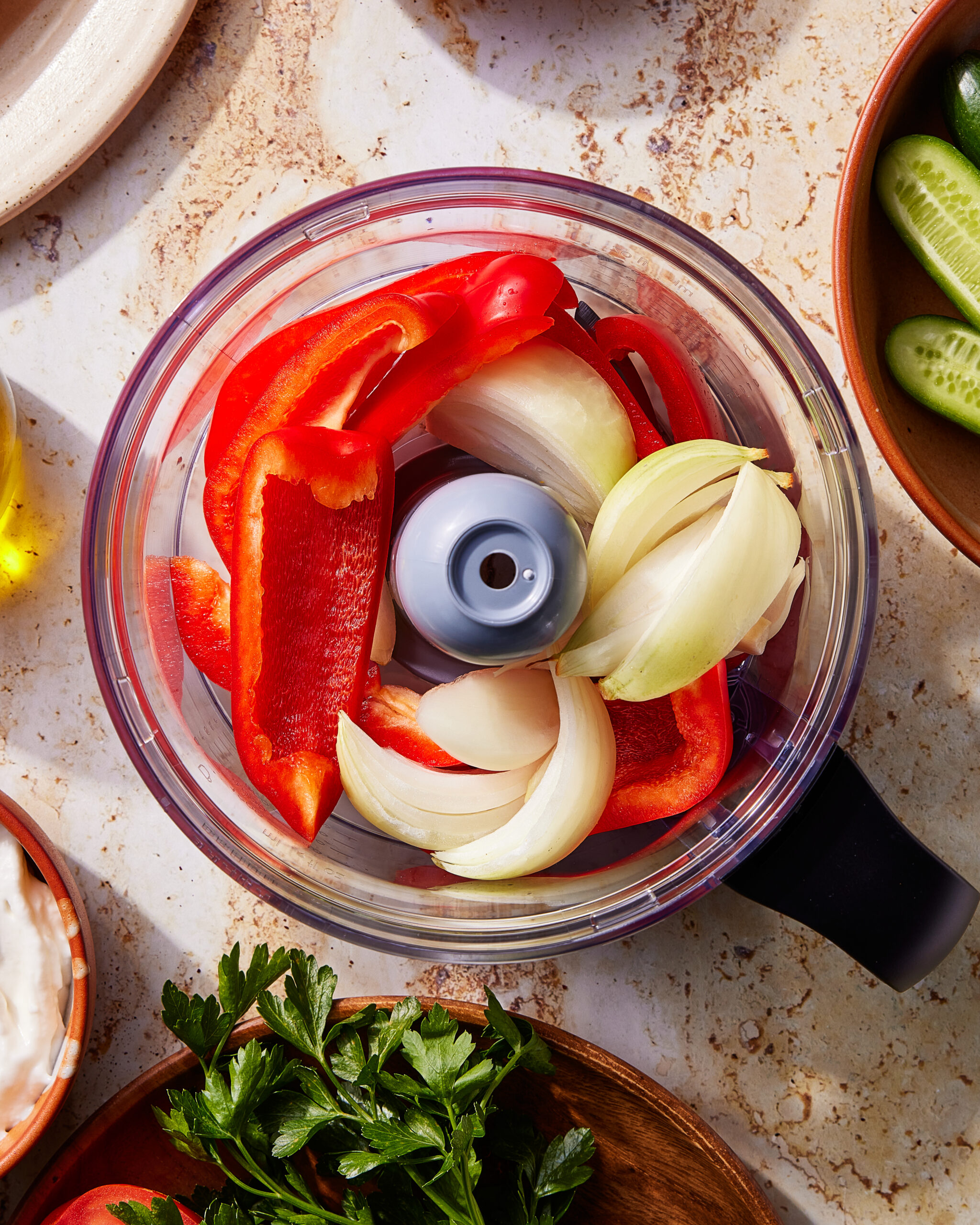 A food processor bowl filled with sliced red bell peppers, onion wedges, and garlic cloves sits on a countertop alongside bowls of cucumbers, herbs, and other ingredients.