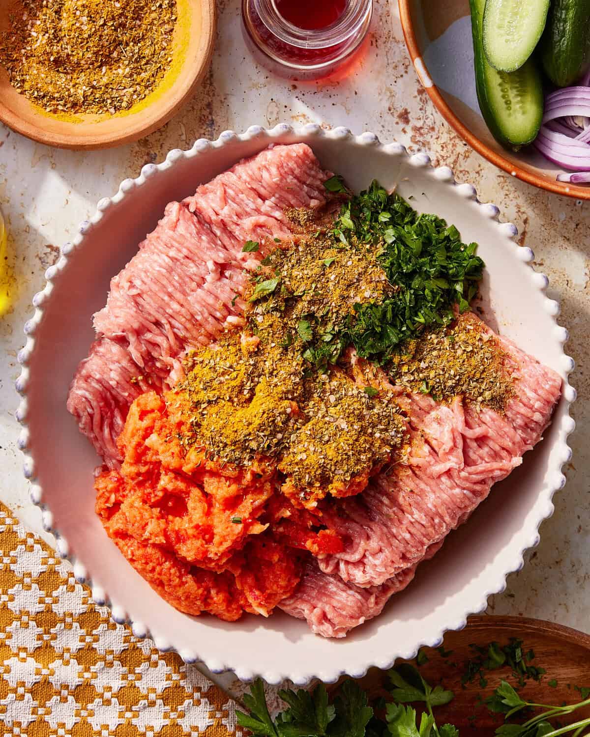 A bowl with raw ground meat, chopped herbs, spices, and a red paste, surrounded by small dishes of spices, cucumber slices, and sliced red onions.