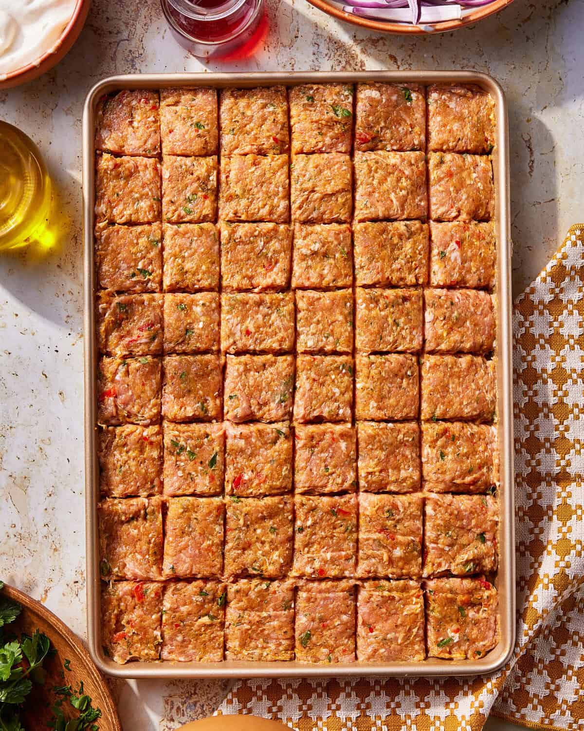 A baking tray filled with raw, seasoned ground meat mixture, evenly scored into square portions. The tray sits on a light marble surface next to a patterned napkin and small bowls of ingredients.