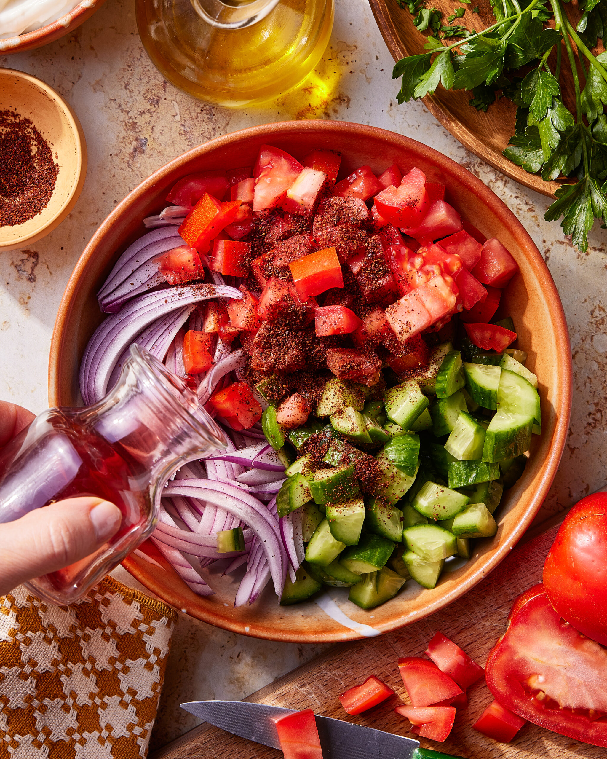 A hand pours oil onto a bowl of sliced red onions, cucumbers, and tomatoes, sprinkled with spices. Fresh parsley, a knife, and more tomatoes are nearby on a countertop.