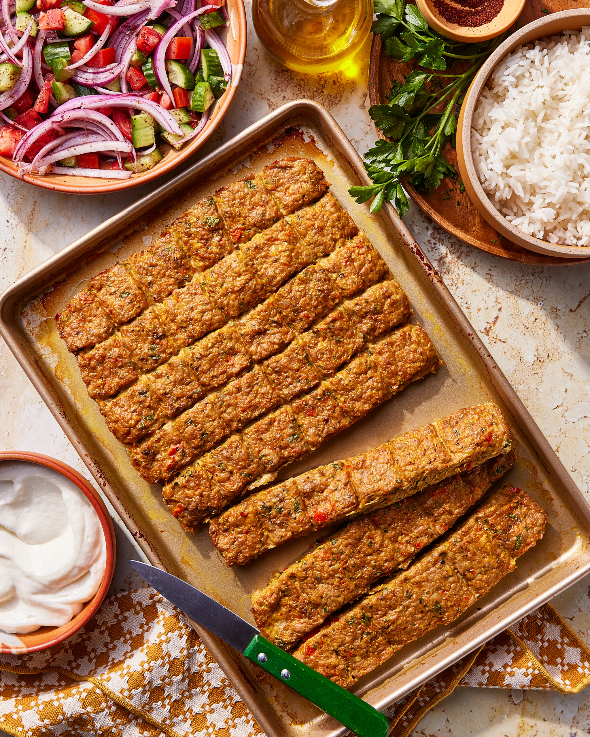 Baked kebab slices on a baking tray with a knife, served alongside a bowl of rice, a bowl of yogurt, a mixed salad, fresh parsley, and a bottle of olive oil.