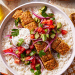 A bowl of white rice topped with grilled kebab slices, chopped cucumber, tomato, red onion, fresh parsley, and a dollop of yogurt sauce, garnished with spices.
