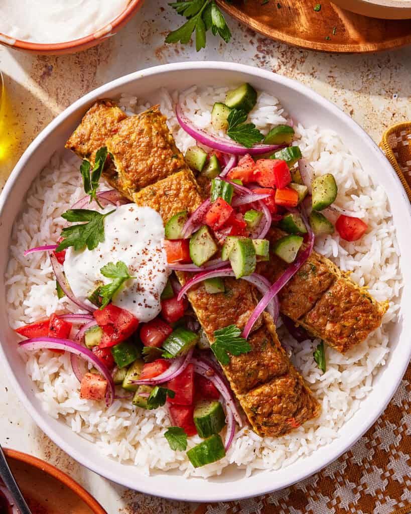 A bowl of white rice topped with grilled kebab slices, chopped cucumber, tomato, red onion, fresh parsley, and a dollop of yogurt sauce, garnished with spices.